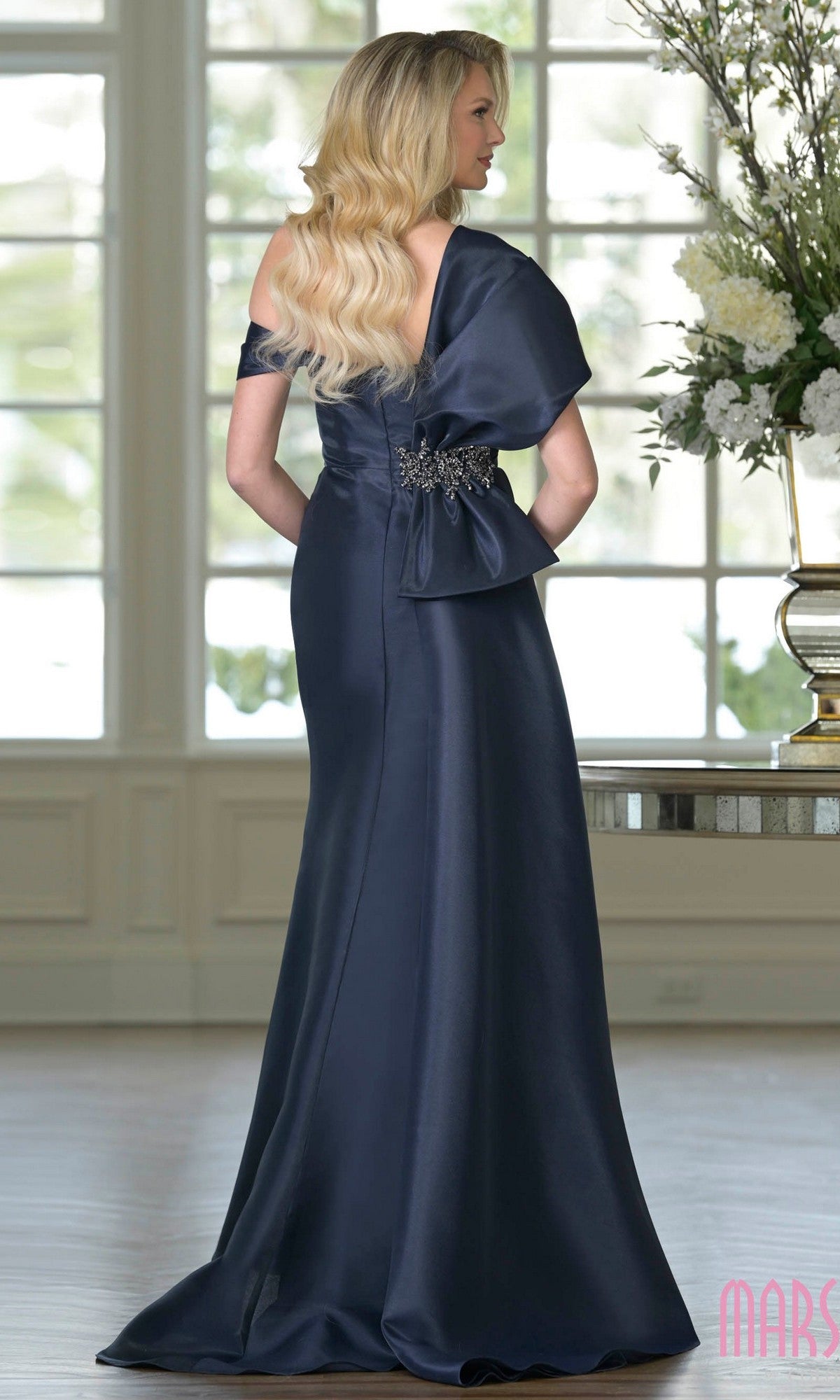 A woman with long blonde hair wears the Marsoni MV1405, a floor-length navy blue formal dress with a bow and beaded applique at the back, standing in an elegant room with large windows and floral arrangements.
